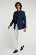 Man wearing a navy blazer, white pants, and navy shoes on a light gray background