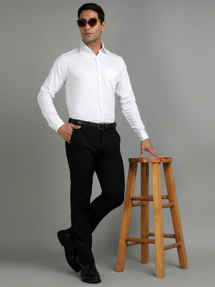Man wearing a white dress shirt and black pants standing next to a wooden stool on a gray background
