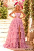 A strapless Rose Pink tiered gown with a high slit, adorned with glittery lace, posed on stone steps with flowering plants.