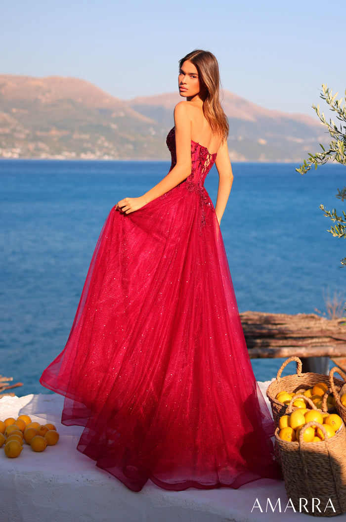 A woman in a sparkly red ball gown stands by the ocean with lemons, mountains in the background.