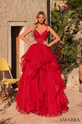 A model wears a dramatic red ball gown with a lace bodice and ruffled tulle skirt in an outdoor Mediterranean setting.