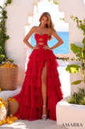 A model wears a flowing red tiered gown with cutouts, standing in a Mediterranean-style setting with lemons and ocean view.